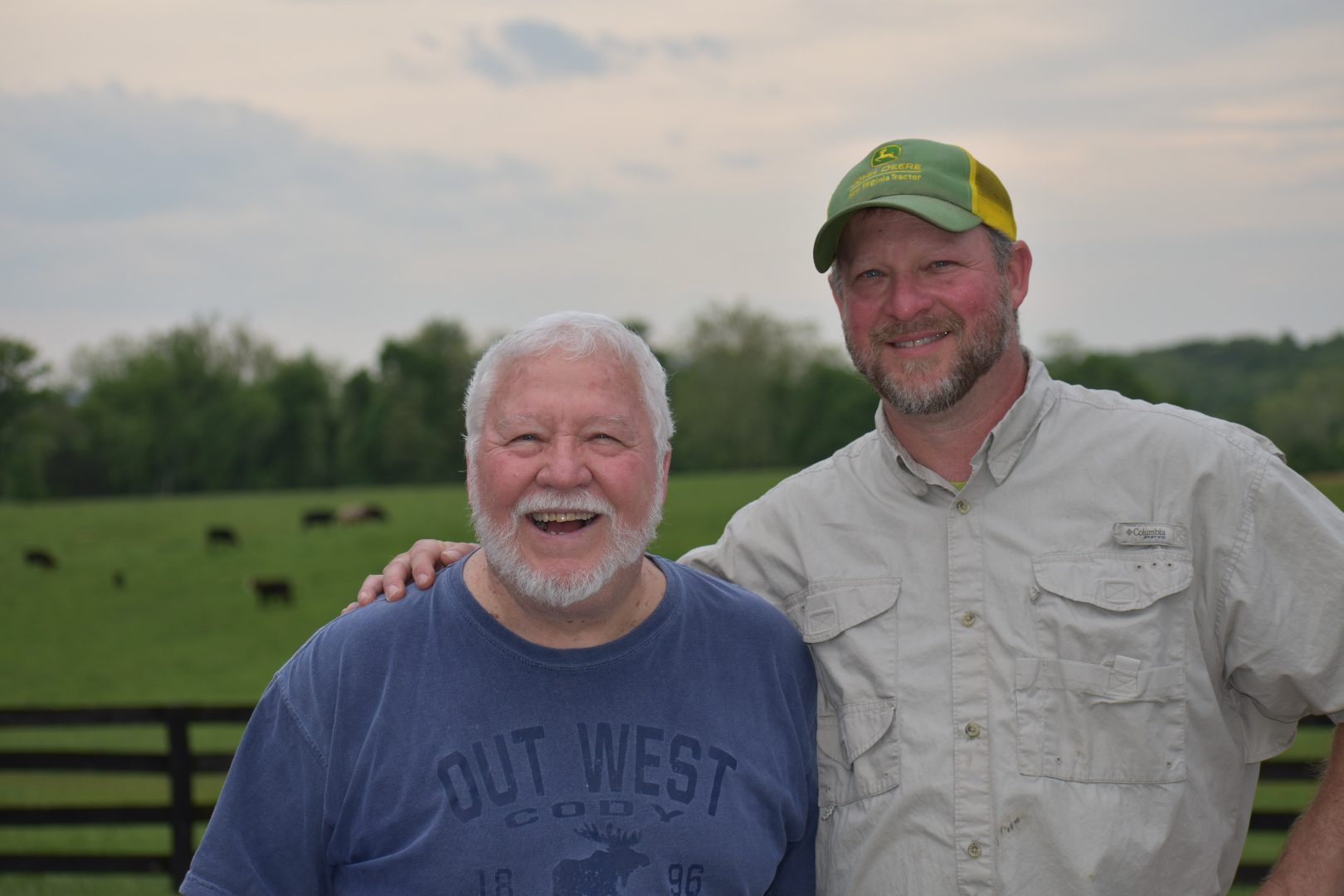 Two men smiling outdoors with cattle in the background.