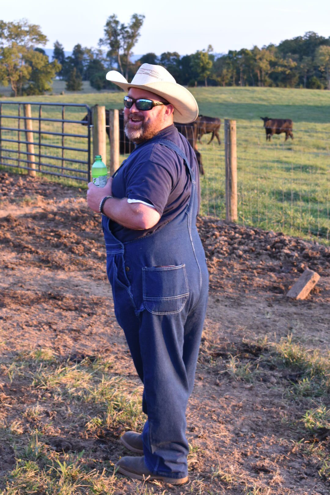 Man in overalls and cowboy hat outdoors near a fenced area.