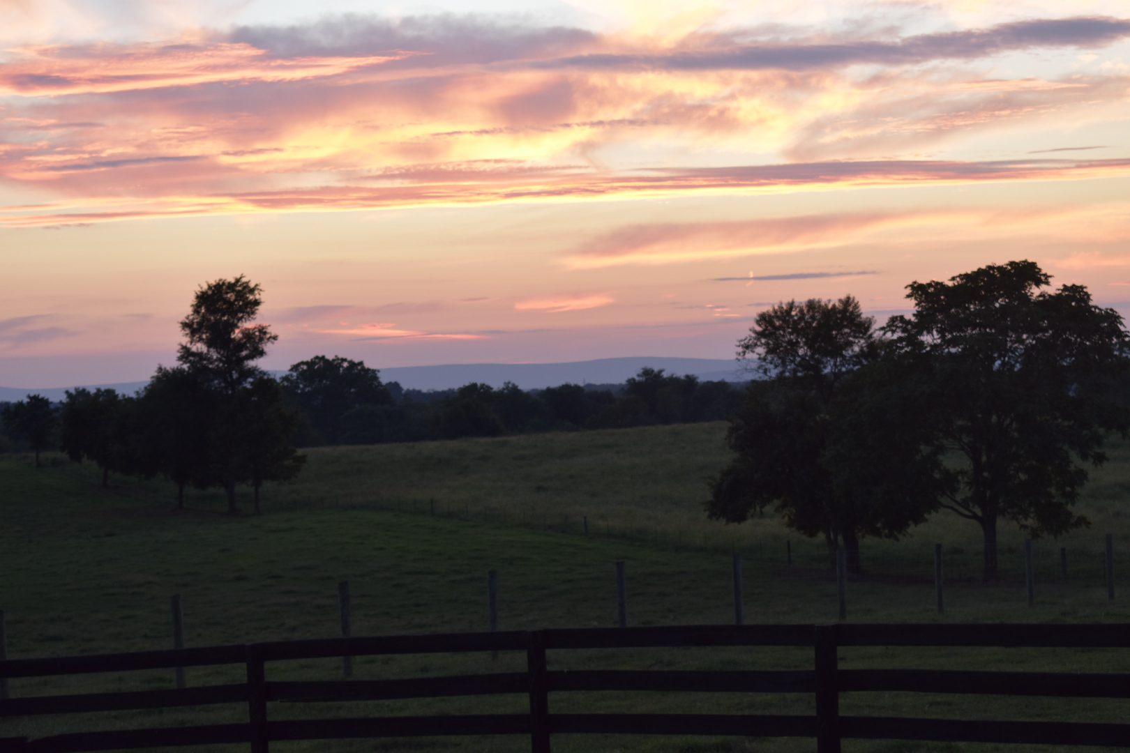 Sunset over a peaceful countryside with silhouetted trees and a wooden fence.