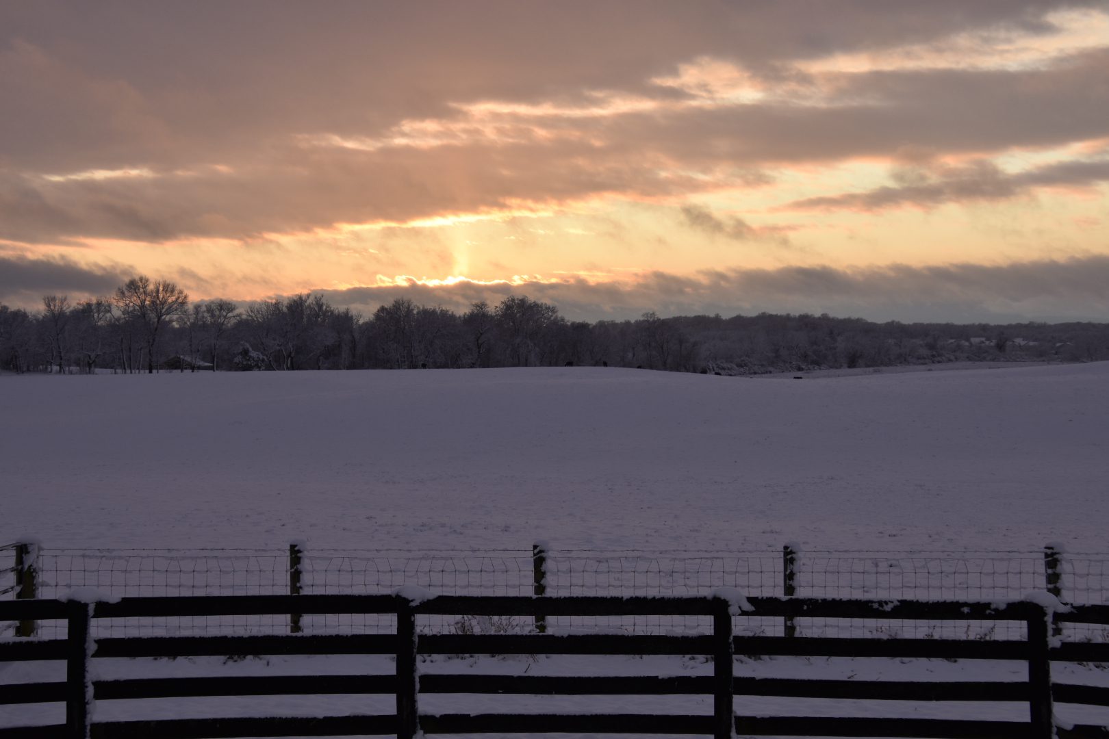 Sunset over a snowy field with a wooden fence in the foreground.